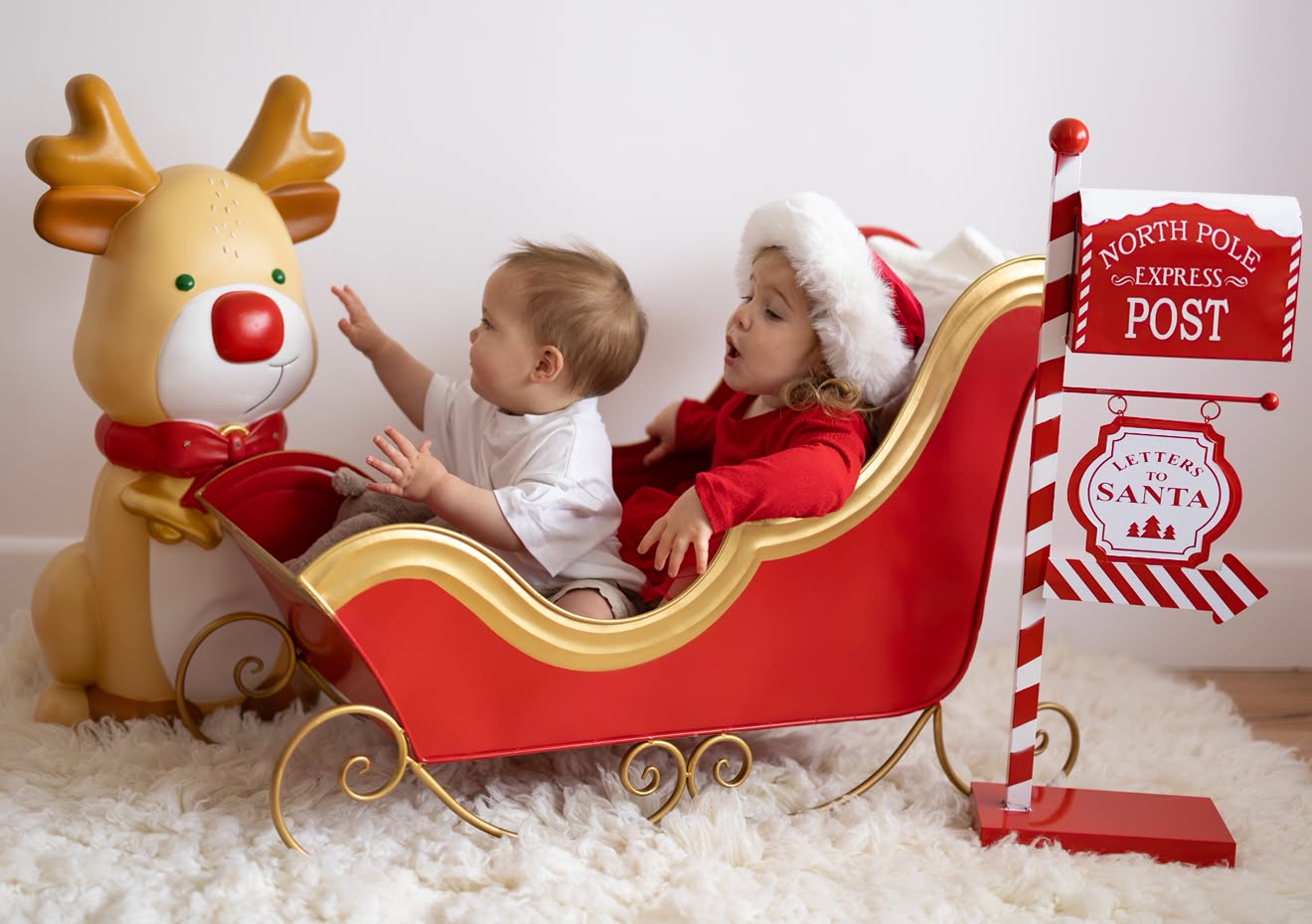 Baby reaching for Rudolph while big sister in a Santa hat sits beside him in a red Christmas sleigh
