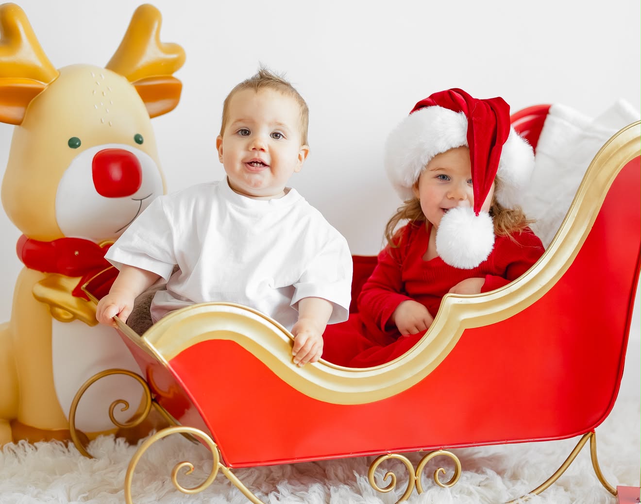 Smiling baby and his sister in a red Christmas sleigh next to Rudolph